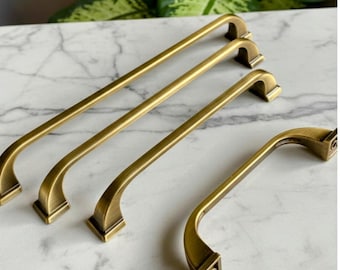 May include: Several antique brass cabinet pulls in different sizes. The pulls have a curved design with square bases, arranged on a marble surface. The brass finish gives them a vintage appearance.