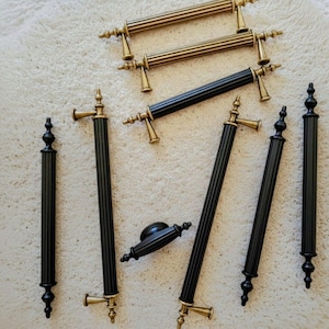 May include: An assortment of cabinet hardware, including handles and pulls. The handles are gold and black, with a ribbed design. The pulls are black with gold accents. The hardware is arranged on a white, textured surface.