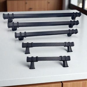 May include: A collection of black cabinet pulls in various sizes. The pulls have a grooved design on the bar and are mounted on a white surface. The hardware is displayed against a backdrop of wooden cabinetry.