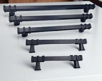 May include: A selection of black cabinet handles in different sizes. The handles feature a grooved design on the bar and are displayed on a white surface. The hardware is set against a backdrop of wooden cabinetry.