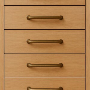 May include: Close-up of a wooden cabinet with five drawers. Each drawer has a horizontal, brass-colored handle. The wood grain is visible, and the cabinet has a light brown color.