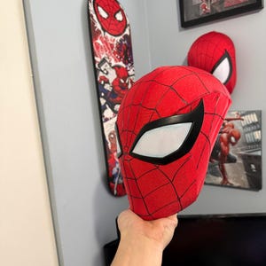 Puede incluir: Una m&aacute;scara roja de Spider-Man con detalles negros y lentes blancos se sostiene. Un monopat&iacute;n y una obra de arte enmarcada con Spider-Man son visibles en el fondo, en una pared gris.