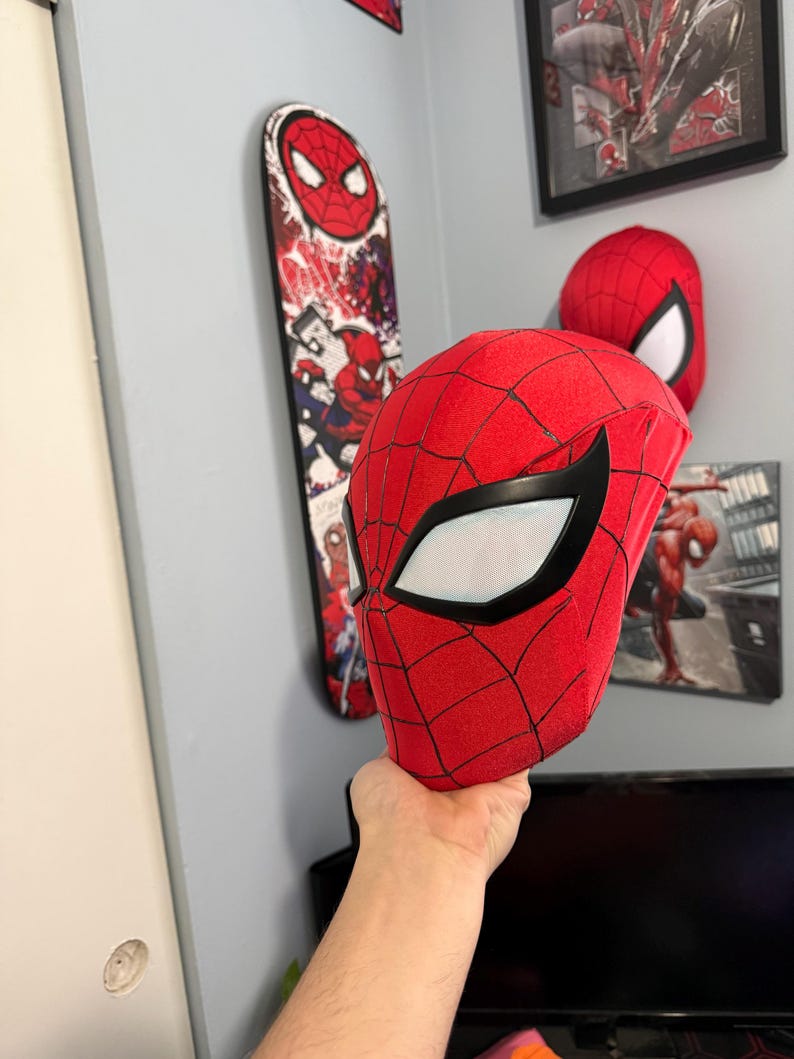 Puede incluir: Una m&aacute;scara roja de Spider-Man con detalles negros y lentes blancos se sostiene. Un monopat&iacute;n y una obra de arte enmarcada con Spider-Man son visibles en el fondo, en una pared gris.