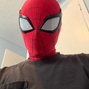 Puede incluir: Una m&aacute;scara roja de Spider-Man con telara&ntilde;as negras y grandes lentes oculares blancas. La m&aacute;scara cubre toda la cabeza y el cuello. La persona lleva una camiseta negra.