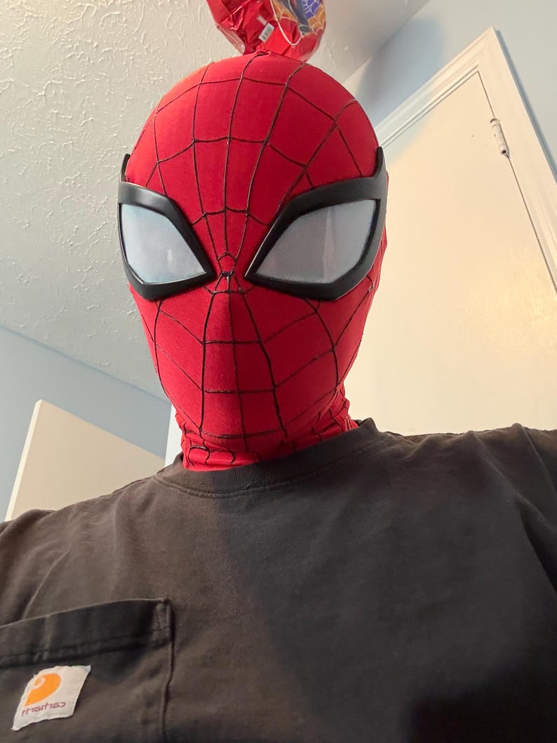 Puede incluir: Una m&aacute;scara roja de Spider-Man con telara&ntilde;as negras y grandes lentes oculares blancas. La m&aacute;scara cubre toda la cabeza y el cuello. La persona lleva una camiseta negra.