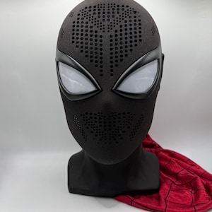 Könnte beinhalten: Eine schwarze Vollkopfmaske mit einer texturierten Oberfläche, die zahlreiche kleine kreisförmige Perforationen im Stirn- und Mundbereich aufweist. Die Maske enthält zwei große, mandelförmige, weiß vernetzte Augenlinsen mit schwarzer Umrandung.