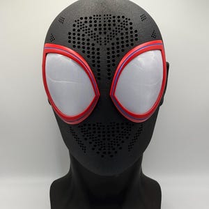 Könnte beinhalten: Eine schwarze Vollkopfmaske mit texturierter Oberfläche und zahlreichen kleinen Belüftungslöchern im Stirn- und Mundbereich. Die Maske verfügt über zwei große, mandelförmige Augenlinsen, die mit einem roten und blauen Rand versehen sind.