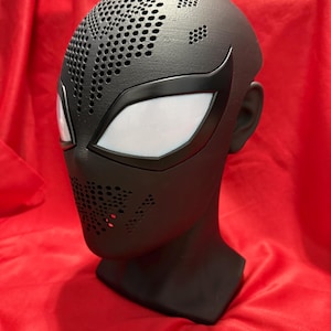 Könnte beinhalten: Eine mattschwarze Vollkopfmaske mit einer texturierten Oberfläche und einem Muster aus kleinen kreisförmigen Perforationen im Stirn- und Mundbereich. Die Maske verfügt über große, kantige weiße Netz-Augenlinsen mit schwarzem Rand.