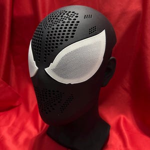 May include: A black mask with white eye shapes, featuring a perforated design. The mask is displayed on a black head form against a red background. The mask is likely a costume or cosplay accessory.
