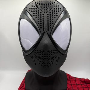 Peut inclure: Un masque intégral noir doté de grands verres oculaires en maille blanche et d'une surface texturée avec des motifs de points perforés sur le front et la bouche, présenté sur un support noir avec une base en tissu à motif toile d'araignée.