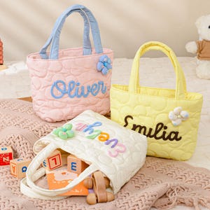 May include: Three quilted tote bags in pastel colours, each embroidered with a name. The pink bag has blue handles and the name "Oliver". The yellow bag has the name "Smilia". The white bag has the name "Evelyn". Toys and a teddy bear are in the background.