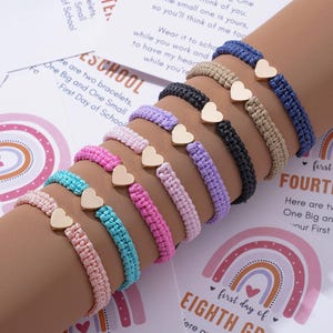 Pode incluir: Uma série de sete pulseiras de cordão trançado em várias cores, incluindo azul, bege, preto, lavanda, rosa claro, rosa choque e turquesa. Cada pulseira apresenta um pequeno pingente de coração de metal dourado polido no centro.