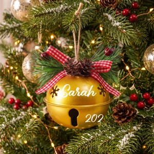May include: A gold-colored metal jingle bell ornament featuring the name Sarah and the number 202 in white text. The bell is decorated with a red and white gingham ribbon, small pinecones, and green pine needles, hanging from a jute twine loop.