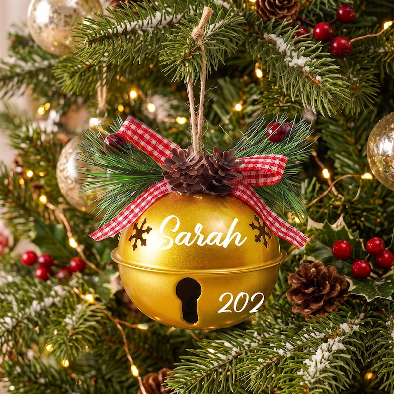 May include: A gold-colored metal jingle bell ornament featuring the name Sarah and the number 202 in white text. The bell is decorated with a red and white gingham ribbon, small pinecones, and green pine needles, hanging from a jute twine loop.