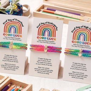 Op de afbeelding: Drie sets van twee gevlochten koordarmbanden op witte displaykaarten. Elke kaart bevat een regenboogafbeelding, de tekst First Day of Preschool, Kindergarten of High School, en een gedicht. De armbanden zijn voorzien van kleine metalen bedels.