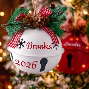 May include: A white metal jingle bell ornament featuring the name Brooks and the year 2026 in red script. The ornament is topped with a pine sprig, a small pinecone, and a red and white checkered ribbon bow.