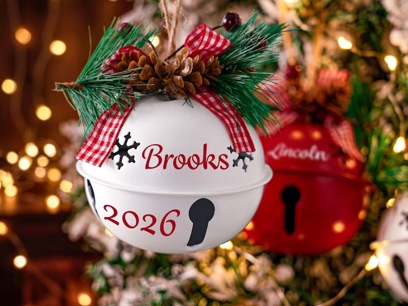 May include: A white metal jingle bell ornament featuring the name Brooks and the year 2026 in red script. The ornament is topped with a pine sprig, a small pinecone, and a red and white checkered ribbon bow.