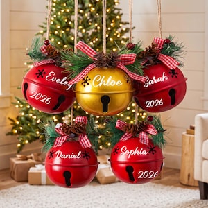 May include: Five round metal jingle bell ornaments hanging by twine. Four are red and one is gold. Each features a pine sprig, pinecone, and red-and-white checkered bow. They are personalized with names and the year 2026 in white text.