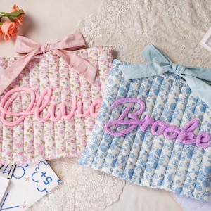 May include: Two rectangular quilted fabric pouches featuring floral patterns. The left pouch is pink with a pink bow and the name Olivia in pink script. The right pouch is blue with a light blue bow and the name Brooke in purple script.