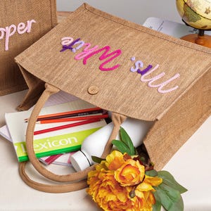 May include: A rectangular brown jute tote bag featuring the embroidered text "Mrs. White" in pink and purple script on the front. The bag has two matching jute handles, a small wooden button closure, and contains a white bottle, pencils, and a book.