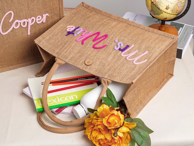 May include: A rectangular brown jute tote bag featuring the embroidered text "Mrs. White" in pink and purple script on the front. The bag has two matching jute handles, a small wooden button closure, and contains a white bottle, pencils, and a book.