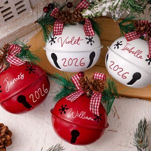 May include: Four round metal jingle bell ornaments in red and white, each personalized with a name and the year 2026. The bells feature red and white checkered bows, pine needles, and pinecones, with black snowflake accents.