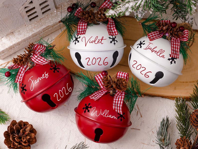 May include: Four round metal jingle bell ornaments in red and white, each personalized with a name and the year 2026. The bells feature red and white checkered bows, pine needles, and pinecones, with black snowflake accents.