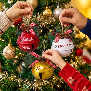 May include: Three round metal bell ornaments in red, white, and gold. Each bell features a name in script font, including Sarch, Lucas, and Kevin, with the year 2026 on the red bell. They are decorated with pine needles, pinecones, and red plaid ribbon.
