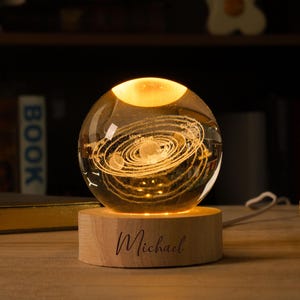 以下が含まれることがあります： 木製の台座に置かれた、太陽系のデザインが施された透明なガラス球。台座には「Michael」という名前が刻まれています。球体は内側から照らされ、温かい光を放っています。装飾品です。