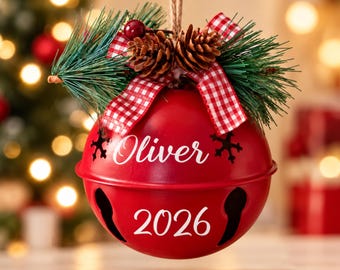 Puede incluir: Un adorno de campana de metal rojo con el nombre Oliver y el año 2026 en letras blancas. La parte superior está decorada con agujas de pino verdes, dos piñas marrones, una baya roja y una cinta de cuadros rojos y blancos.