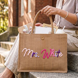 May include: A rectangular tan jute tote bag featuring a single wooden button closure at the top centre. The front of the bag displays the text Mrs. White in raised, cursive-style felt lettering in shades of light pink, dark pink, and purple.