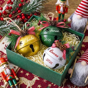 May include: Three round metal jingle bell ornaments in gold, green, and white, each personalized with a name and the year 2026. Each bell is topped with a red and white checkered ribbon, pine needles, and small pinecones, resting in a green box.