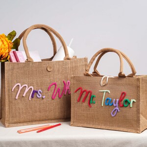 May include: Two rectangular hessian tote bags with brown handles and a single button closure. The larger bag features the pink and purple embroidered text Mrs. Wh, while the smaller bag displays the red, green, and purple embroidered text Mr. Taylor.