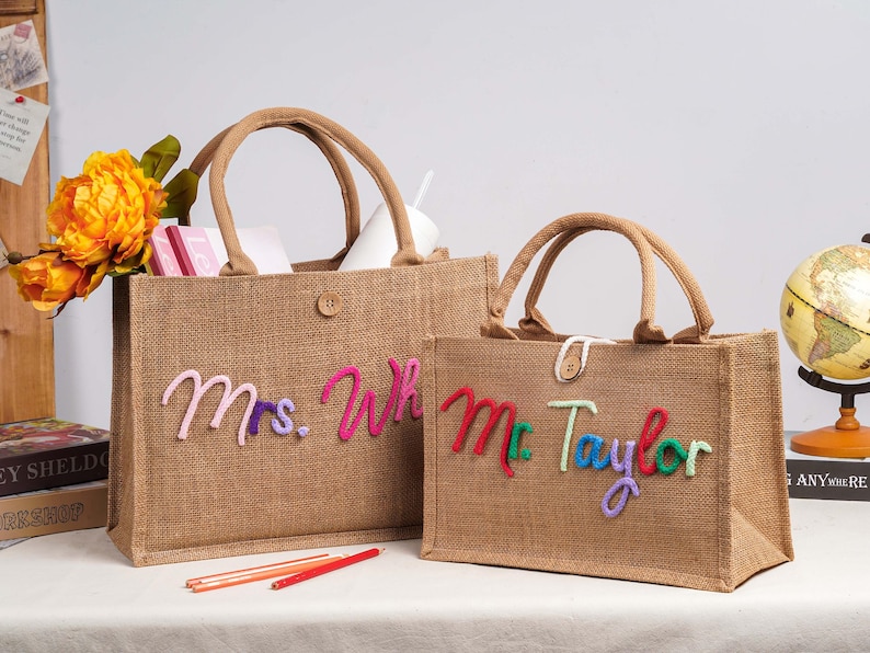 May include: Two rectangular hessian tote bags with brown handles and a single button closure. The larger bag features the pink and purple embroidered text Mrs. Wh, while the smaller bag displays the red, green, and purple embroidered text Mr. Taylor.