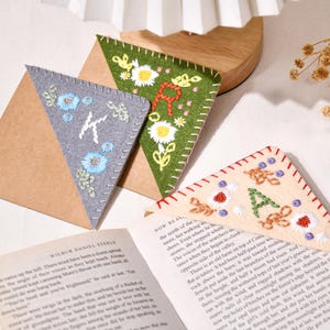 以下が含まれることがあります： グレー、グリーン、ベージュの刺繍フェルトブックマーク。それぞれ異なるイニシャルと花の模様が付いています。ブックマークは本の開いたページに配置されています。ブックマークの幅は約7.6cmです。