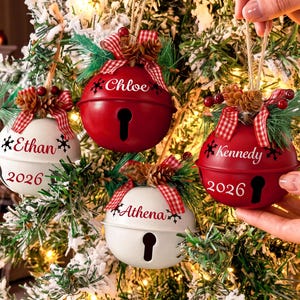 May include: Four round metal jingle bell ornaments in red and white, each personalized with a name and the year 2026. The ornaments feature a red and white checkered bow, pine needles, and small pinecones at the top with a keyhole opening.