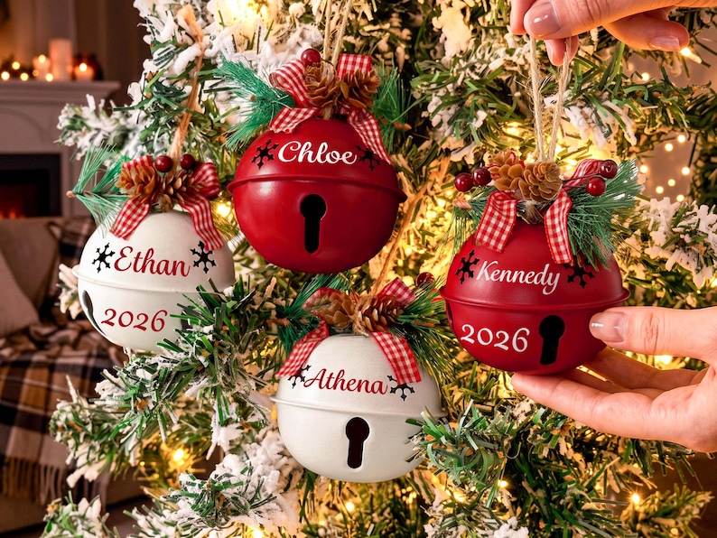 May include: Four round metal jingle bell ornaments in red and white, each personalized with a name and the year 2026. The ornaments feature a red and white checkered bow, pine needles, and small pinecones at the top with a keyhole opening.