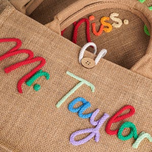 May include: A tan hessian tote bag featuring the text Mr. Taylor in raised, multi-coloured rope lettering. The bag includes a brown wooden button closure with a white rope loop. The texture of the woven fabric is visible throughout the surface.