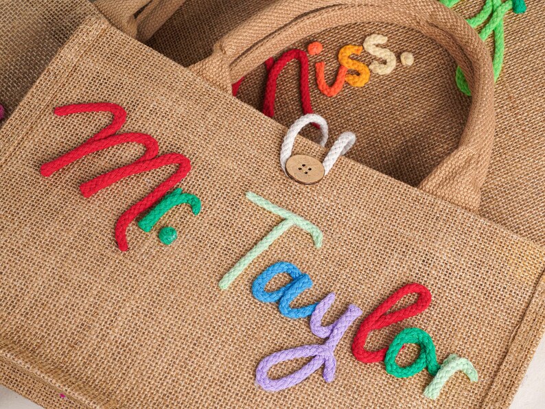 May include: A tan hessian tote bag featuring the text Mr. Taylor in raised, multi-coloured rope lettering. The bag includes a brown wooden button closure with a white rope loop. The texture of the woven fabric is visible throughout the surface.