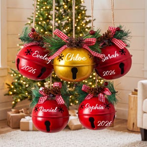 May include: Five hanging jingle bell ornaments, four red and one gold, each personalised with a name and the year 2026. Each bell features a red and white chequered ribbon, pine needles, and a small pinecone attached to the top hanging twine.