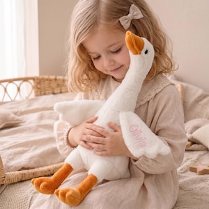 Può includere: Un peluche di oca bianca con zampe e becco arancioni, tenuto in braccio da una bambina. L'oca ha il nome "Olivia" ricamato sull'ala. Il giocattolo è morbido e coccoloso, perfetto per il comfort di un bambino.