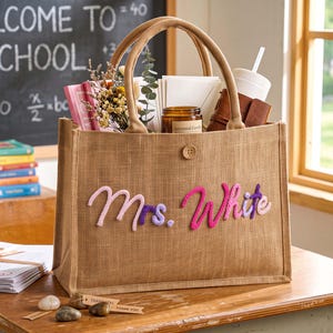 May include: A rectangular tan jute tote bag with two top handles and a single button closure. The front features the name Mrs. White in raised, multi-coloured cursive lettering in shades of pink, purple, and lavender. The bag is filled with items.
