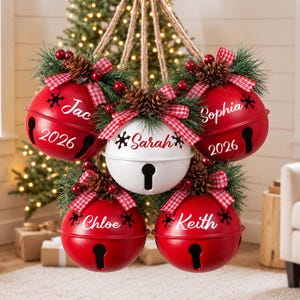 May include: A cluster of five hanging jingle bell ornaments suspended by jute rope. Four red bells and one white bell are decorated with pine needles, pinecones, red berries, and red-and-white checkered bows. Each bell features a name and the year 2026.