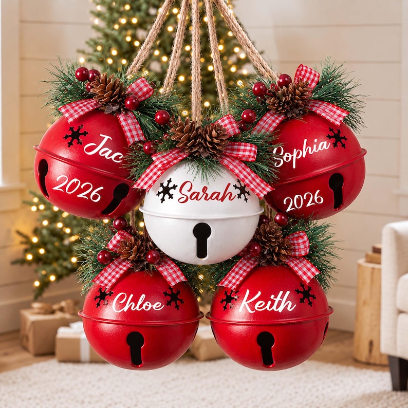 May include: A cluster of five hanging jingle bell ornaments suspended by jute rope. Four red bells and one white bell are decorated with pine needles, pinecones, red berries, and red-and-white checkered bows. Each bell features a name and the year 2026.