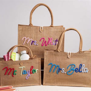 May include: Three rectangular tan hessian tote bags with two handles and a single button closure. Each bag features custom cursive text in colourful yarn: Mrs. White, Mr. Taylor, and Mrs. Bella. The bags are structured with flat bottoms and open tops.