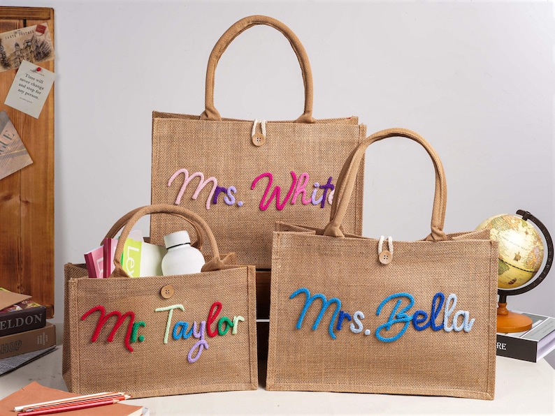 May include: Three rectangular tan hessian tote bags with two handles and a single button closure. Each bag features custom cursive text in colourful yarn: Mrs. White, Mr. Taylor, and Mrs. Bella. The bags are structured with flat bottoms and open tops.