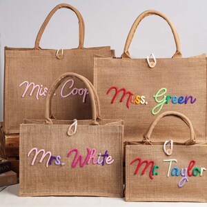 May include: Four rectangular tan jute tote bags with two top handles and a single button closure. Each bag features colourful, raised, cursive embroidered text reading "Mrs. Coop," "Miss. Green," "Mrs. White," and "Mr. Taylor" on the front.
