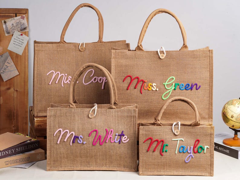 May include: Four rectangular tan jute tote bags with two top handles and a single button closure. Each bag features colourful, raised, cursive embroidered text reading "Mrs. Coop," "Miss. Green," "Mrs. White," and "Mr. Taylor" on the front.