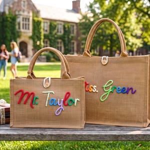 May include: Two rectangular tan jute tote bags with top handles and wooden button closures. The smaller bag features the text Mr. Taylor in colourful raised lettering, while the larger bag displays the text Miss. Green in similar colourful raised lettering.