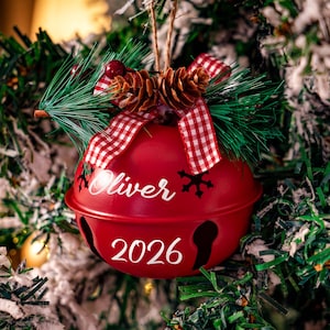 May include: A red metal jingle bell ornament featuring the name Oliver and the year 2026 in white script. The top is decorated with a red and white checkered ribbon, two small pinecones, green pine needles, and red berries.